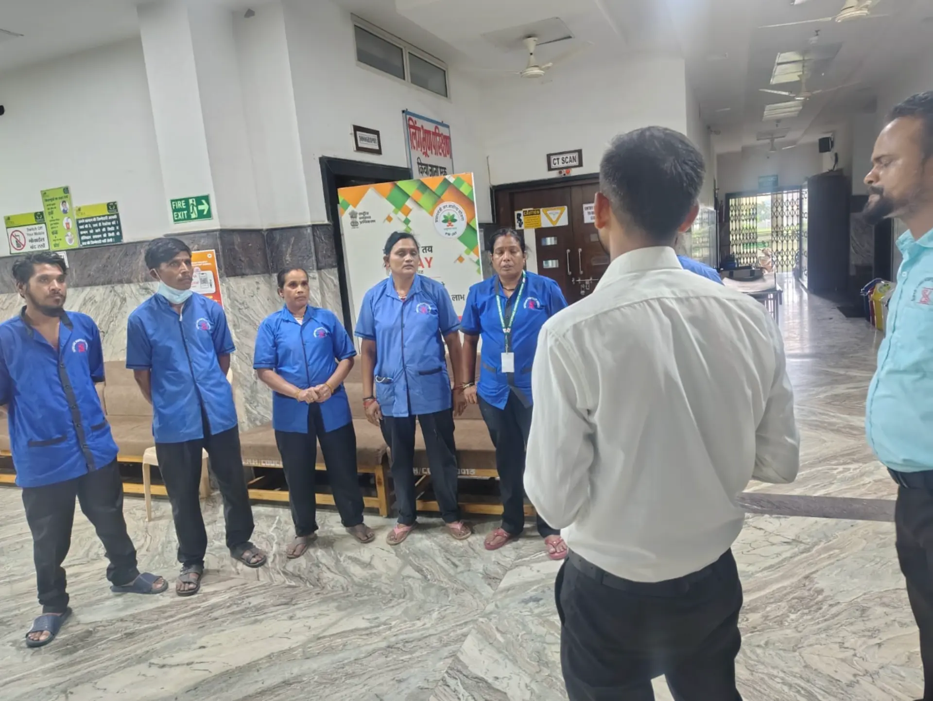 Supervisor conducting a shift briefing with blue-uniformed staff in a hospital lobby